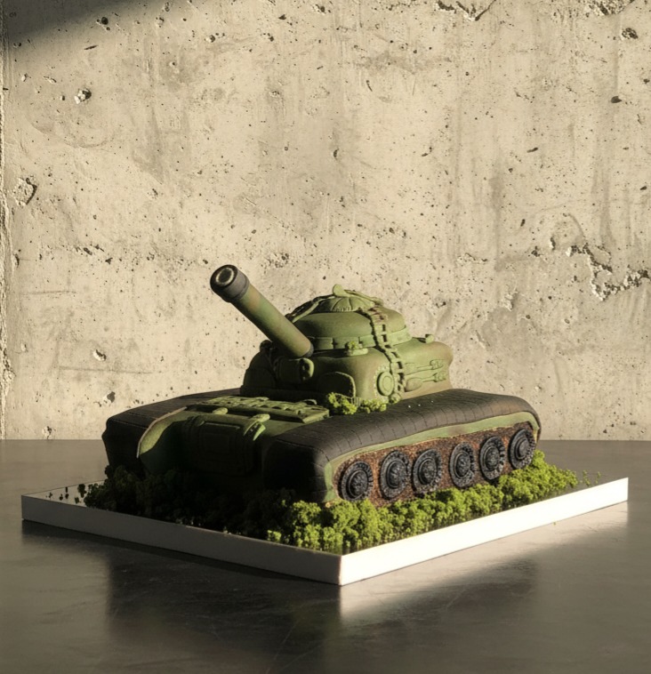 Tort w kształcie czołgu T-72 w odcieniach zieleni, umieszczony na białej podstawie z dekoracją imitującą trawę, na tle surowej, betonowej ściany.