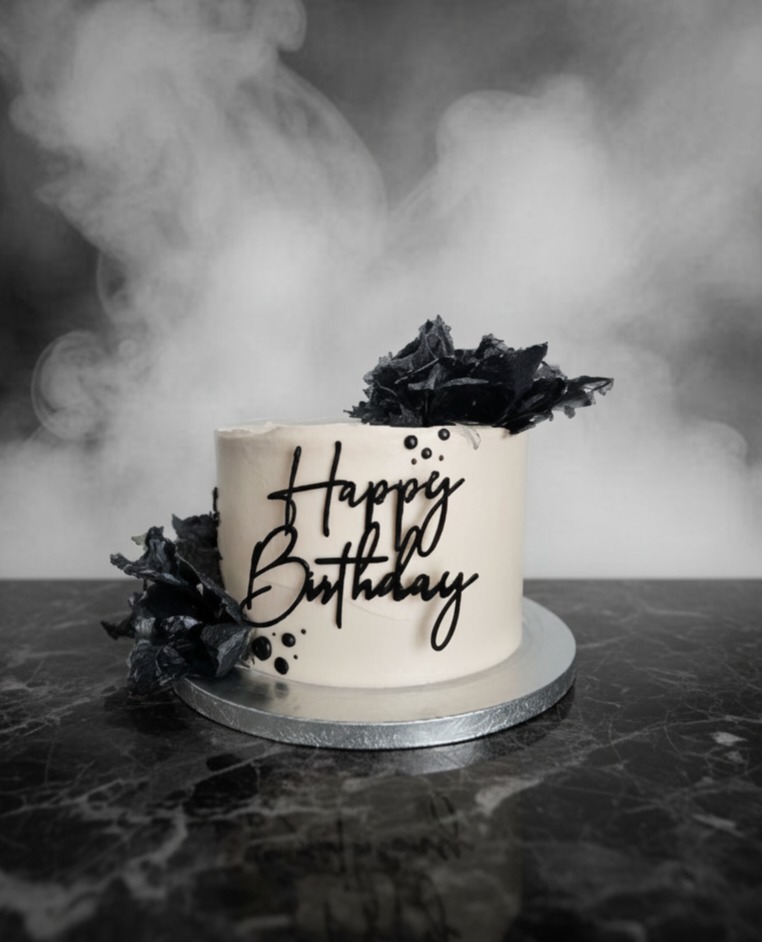 Monochromatyczny tort urodzinowy z czarnym napisem 'Happy Birthday' i dekoracjami z ciemnej czekolady na marmurowym blacie, tło w odcieniach szarości.