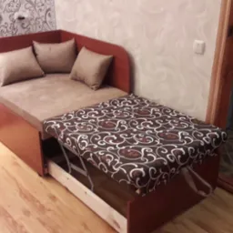 Rozkładana sofa narożna z brązową tapicerką i wzorzystym pokrowcem na wysuwanej części, uzupełniona poduszkami, na tle wzorzystej tapety.