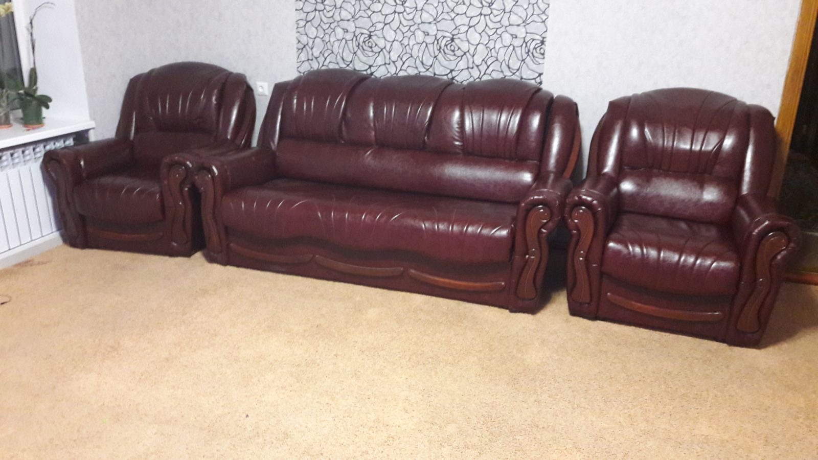 Komplet wypoczynkowy w odcieniach bordo: sofa trzyosobowa i dwa fotele z drewnianymi zdobieniami na tle ściany z wzorem kwiatowym.