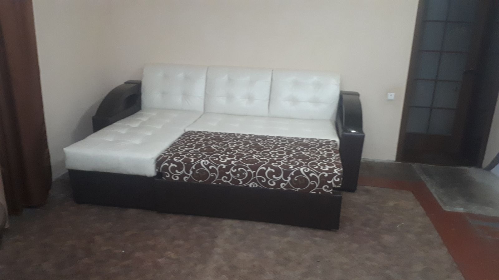 Rozkładany narożnik z białą, pikowaną skórą na górze i wzorzystą tkaniną na dole, stojący na brązowym dywanie w pokoju. Widoczny fragment zasłony i drzwi.