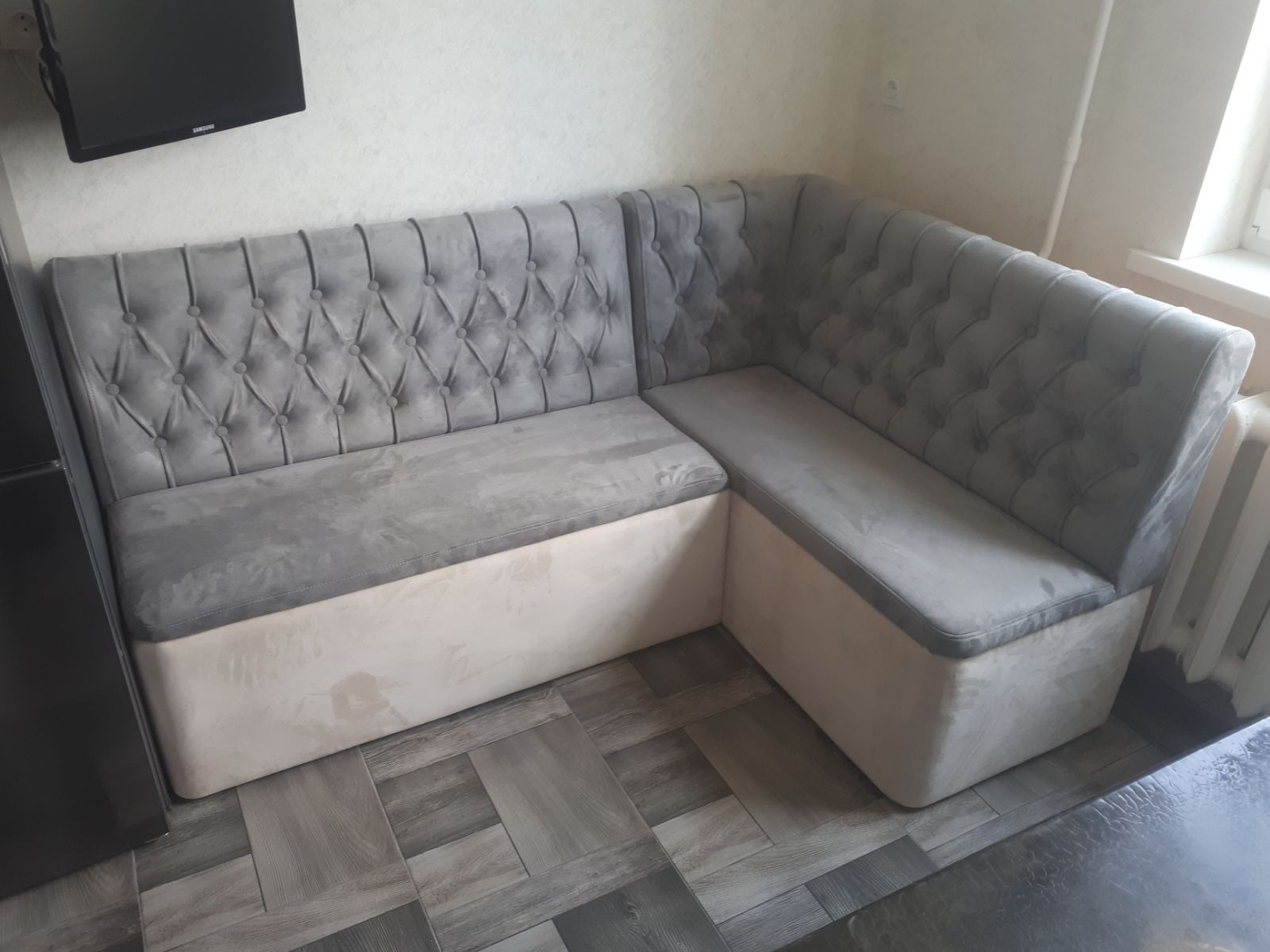 Szary, tapicerowany narożnik kuchenny z pikowanym oparciem i jasną podstawą, stojący na podłodze imitującej drewno, z widocznym fragmentem okna.