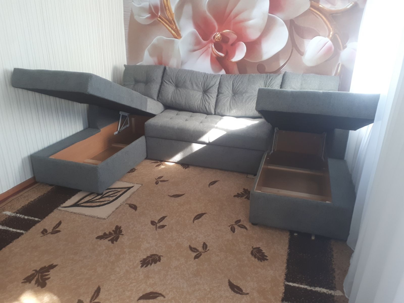 Szary narożnik z otwieranymi schowkami na tle ściany z kwiatowym motywem, widoczny na tle dywanu w Wrześni. Jasne, naturalne światło.