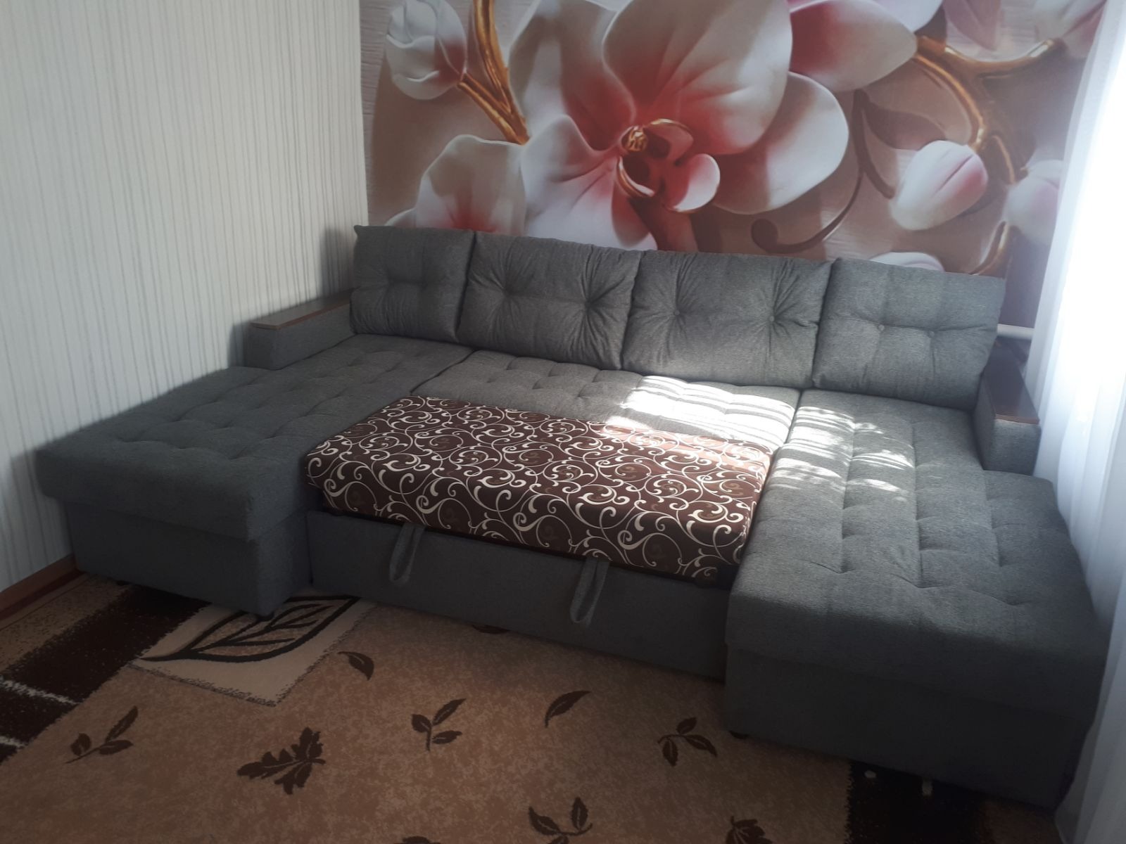 Szary narożnik z brązowym wzorzystym pokrowcem na wysuwanym elemencie, na tle ściany z tapetą w kwiaty i zasłony.