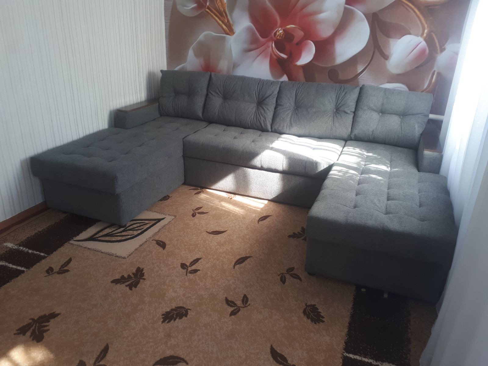 Szary narożnik z pikowanymi poduchami na tle ściany z kwiatowym wzorem. Widoczny dywan z motywem liści. Jasne światło wpada przez okno.