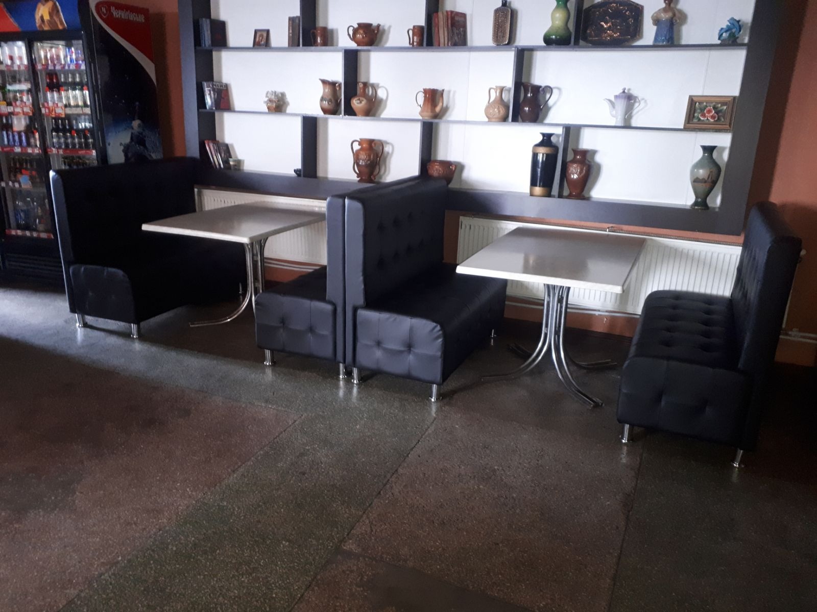 Czarne, tapicerowane loże z białymi stolikami w lokalu. W tle półki z dekoracyjnymi naczyniami. Widok na podłogę z szarego lastryko i dystrybutor napojów.