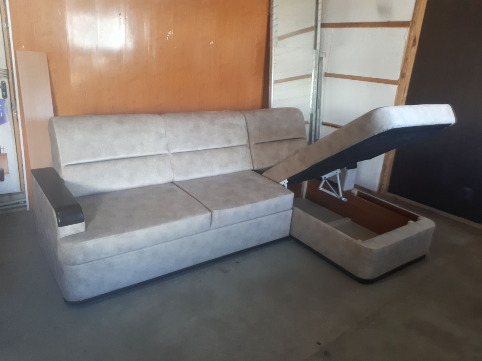 Beżowa sofa narożna z podnoszonym siedziskiem, ukazującym schowek. Mebel stoi na szarym tle, ściany w odcieniach brązu i bieli.