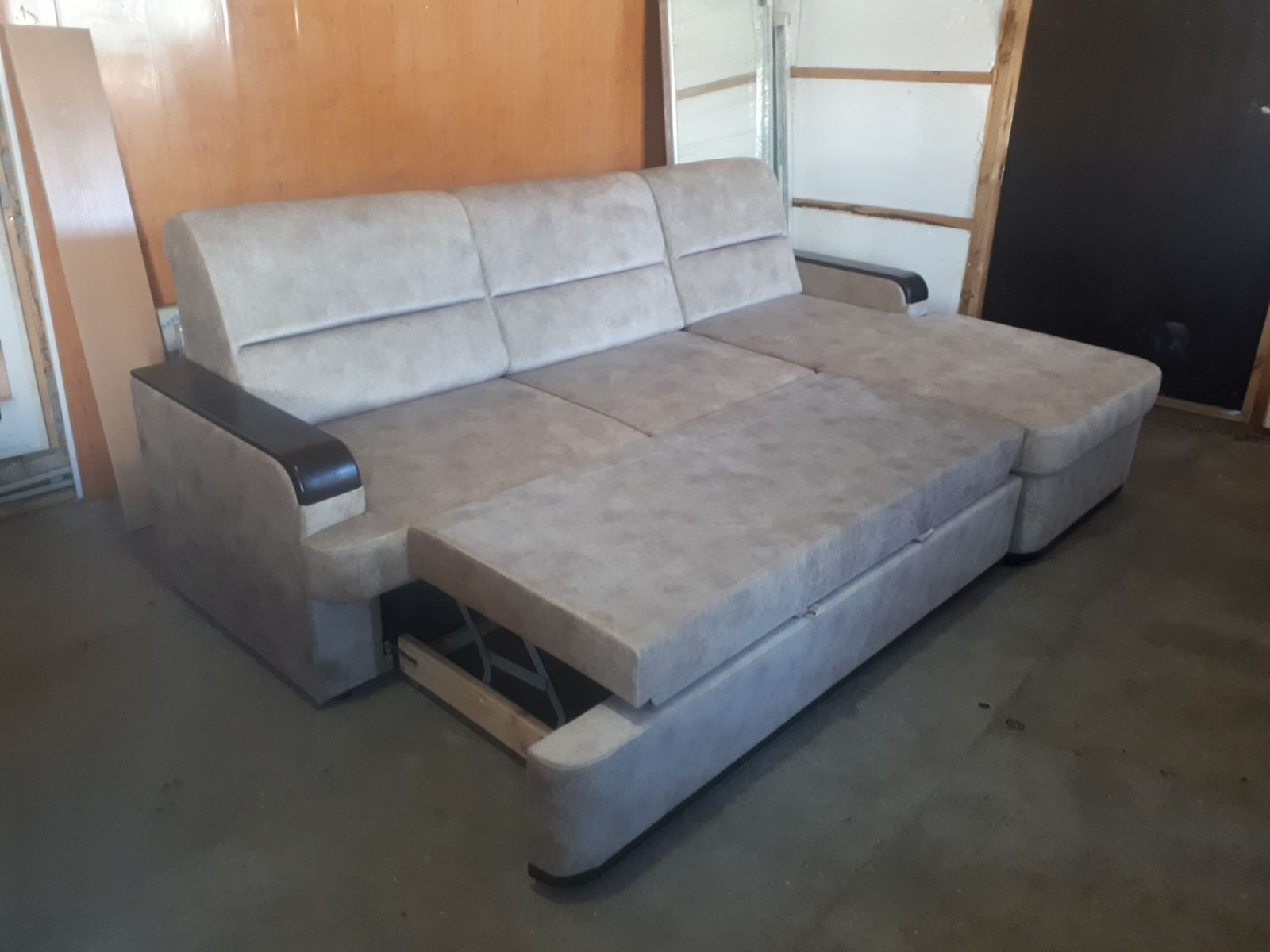 Rozłożona, jasna sofa z ciemnymi podłokietnikami, widoczny mechanizm wysuwania. Ustawiona na tle ściany w pomieszczeniu.