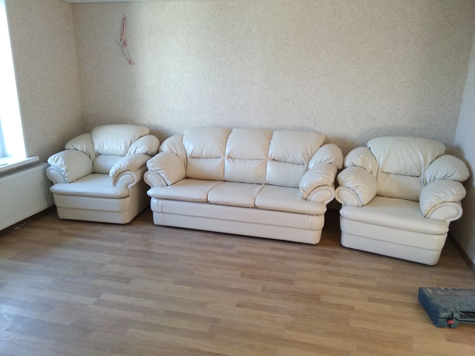 Komplet wypoczynkowy w jasnej skórze: sofa trzyosobowa i dwa fotele z charakterystycznymi, zaokrąglonymi podłokietnikami, ustawione na panelach podłogowych w pokoju.