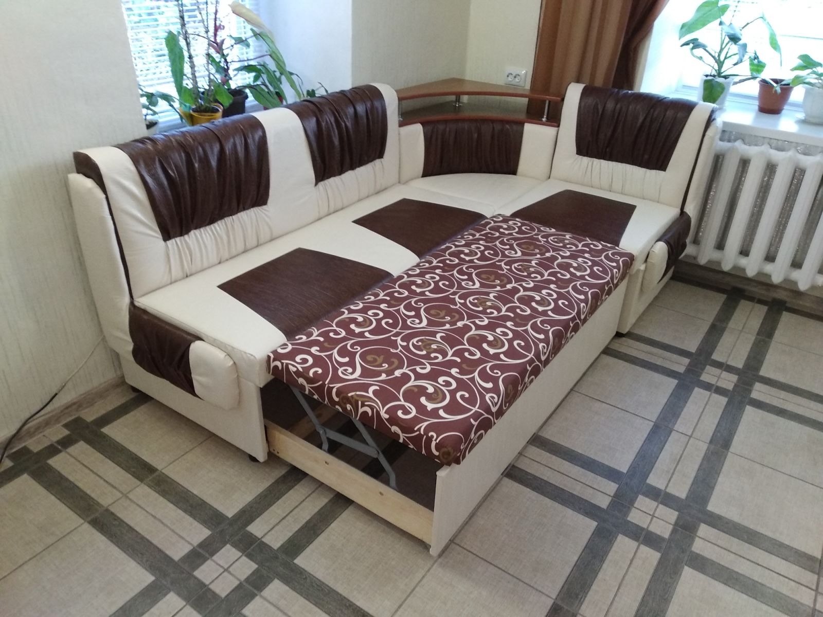 Rozkładana sofa narożna z brązowo-białą tapicerką w kratę, z widocznym mechanizmem rozkładania i wzorzystym pokrowcem na wysuniętym elemencie.