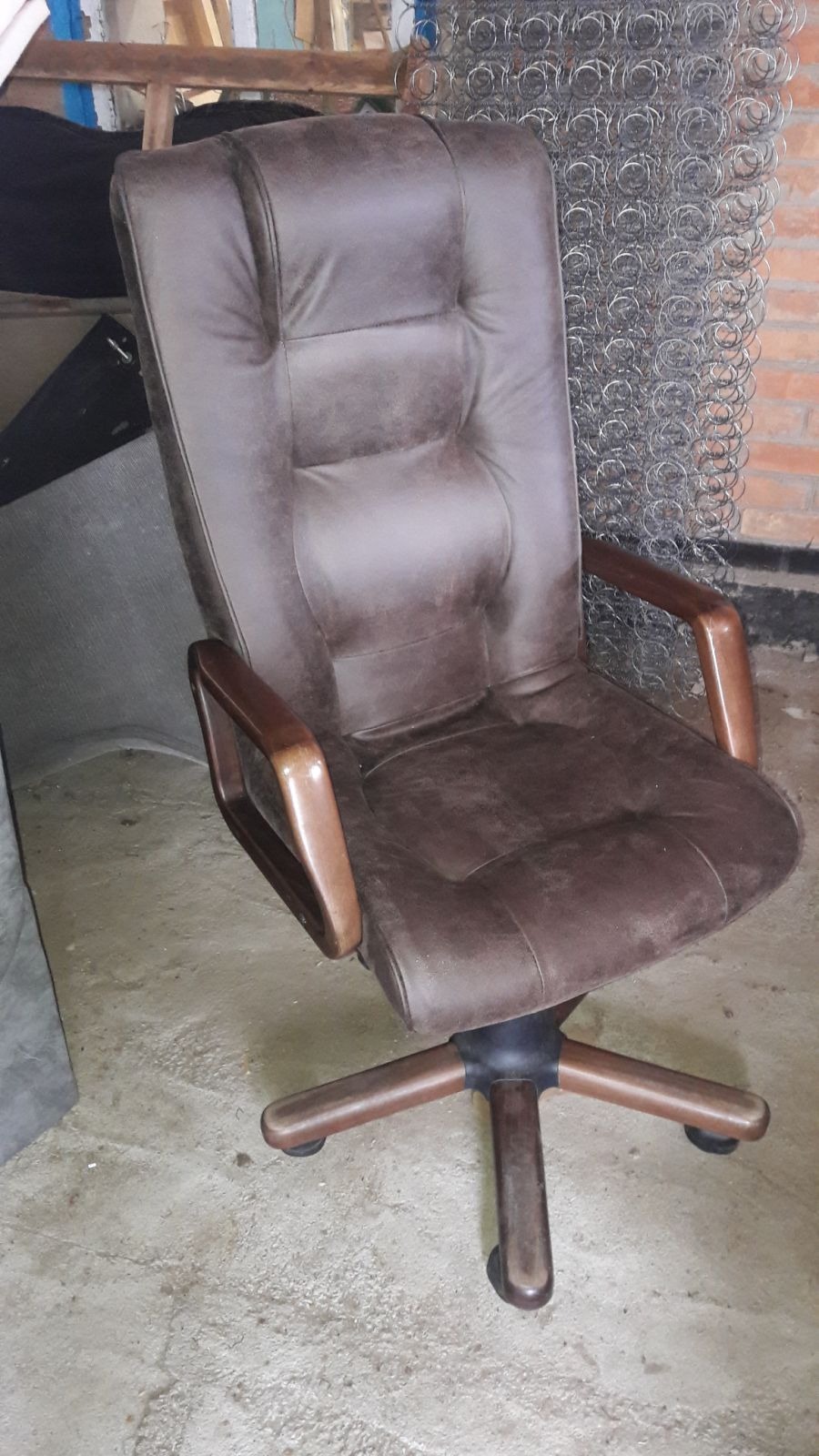 Tapicerowane krzesło biurowe z drewnianymi podłokietnikami, widoczne sprężyny w tle, sugerujące warsztat tapicerski. Brązowa skóra, stan używany, ale zadbany.