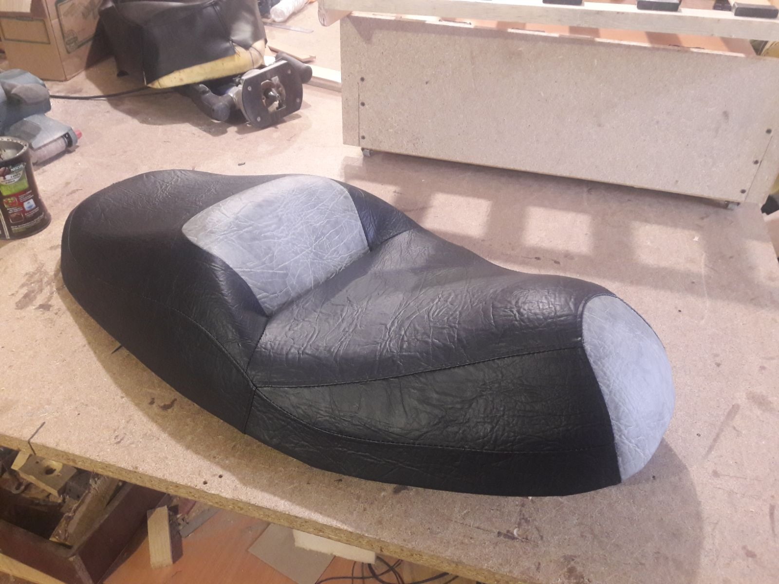 Czarne, skórzane siedzisko motocyklowe z szarymi wstawkami, leżące na warsztatowym stole. Widoczne narzędzia i materiały tapicerskie w tle.