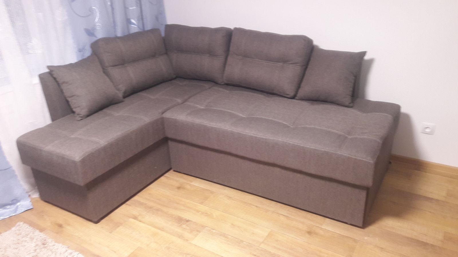 Brązowy narożnik z poduszkami w pokoju, widok z niskiego kąta. Widoczna drewniana podłoga i zasłony w tle. Narożnik z wyraźnymi przeszyciami.