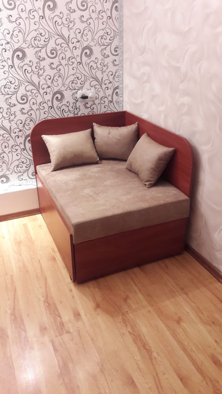 Narożna sofa z beżową tapicerką i trzema poduszkami, ustawiona na drewnianej podłodze w pokoju z wzorzystą tapetą.