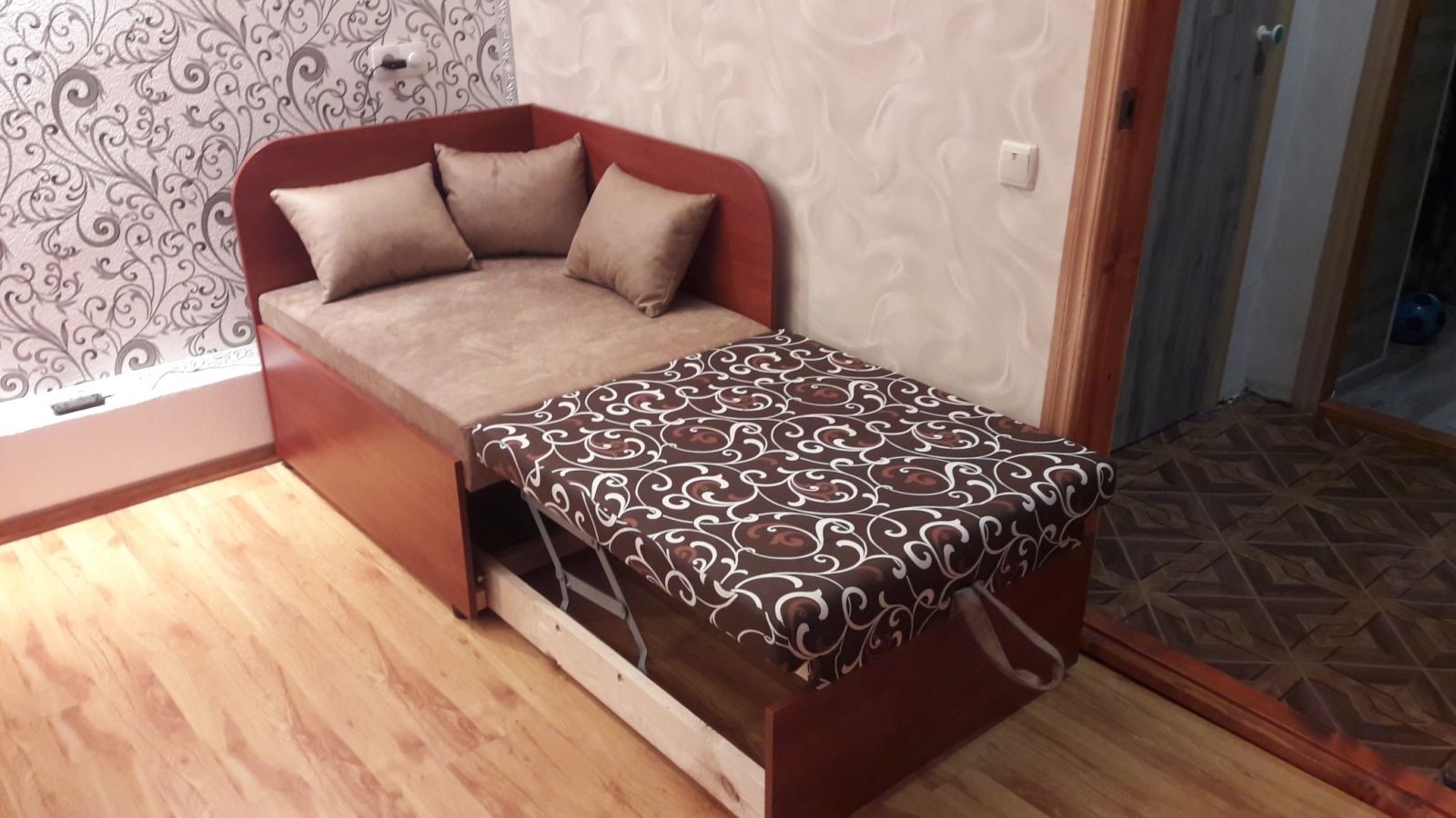 Rozkładana sofa narożna z brązową tapicerką i wzorzystym pokrowcem na wysuwanej części, uzupełniona poduszkami, na tle wzorzystej tapety.