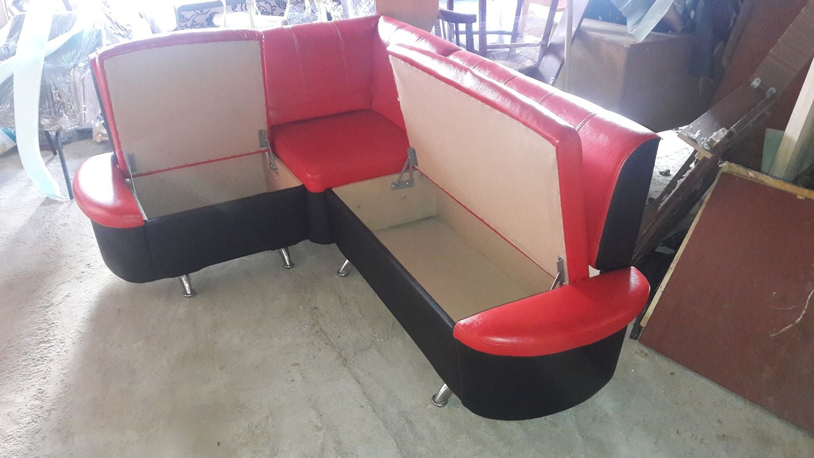 Tapicerowany narożnik w czarno-czerwonej kolorystyce z otwartymi schowkami pod siedziskami, na metalowych nóżkach, w warsztacie.
