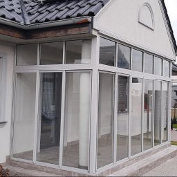 A2U Przemysław Wojciechowski - Szklana zabudowa balkonu w nowoczesnym domu jednorodzinnym z szarymi ramami, widok z zewnątrz na tle ogrodu i sąsiednich budynków.