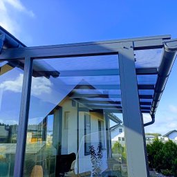 A2U Przemysław Wojciechowski - Szklana zabudowa balkonu z szarymi profilami i widocznymi rynnami, odbijająca otoczenie. Ujęcie z dołu, na tle błękitnego nieba.