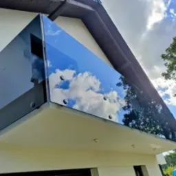 Szklana balustrada balkonowa z widocznym odbiciem chmur na szkle, zamocowana na jasnej elewacji budynku, z drzewem w tle.