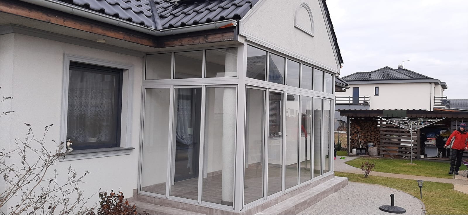 Szklana zabudowa balkonu w nowoczesnym domu jednorodzinnym z szarymi ramami, widok z zewnątrz na tle ogrodu i sąsiednich budynków.