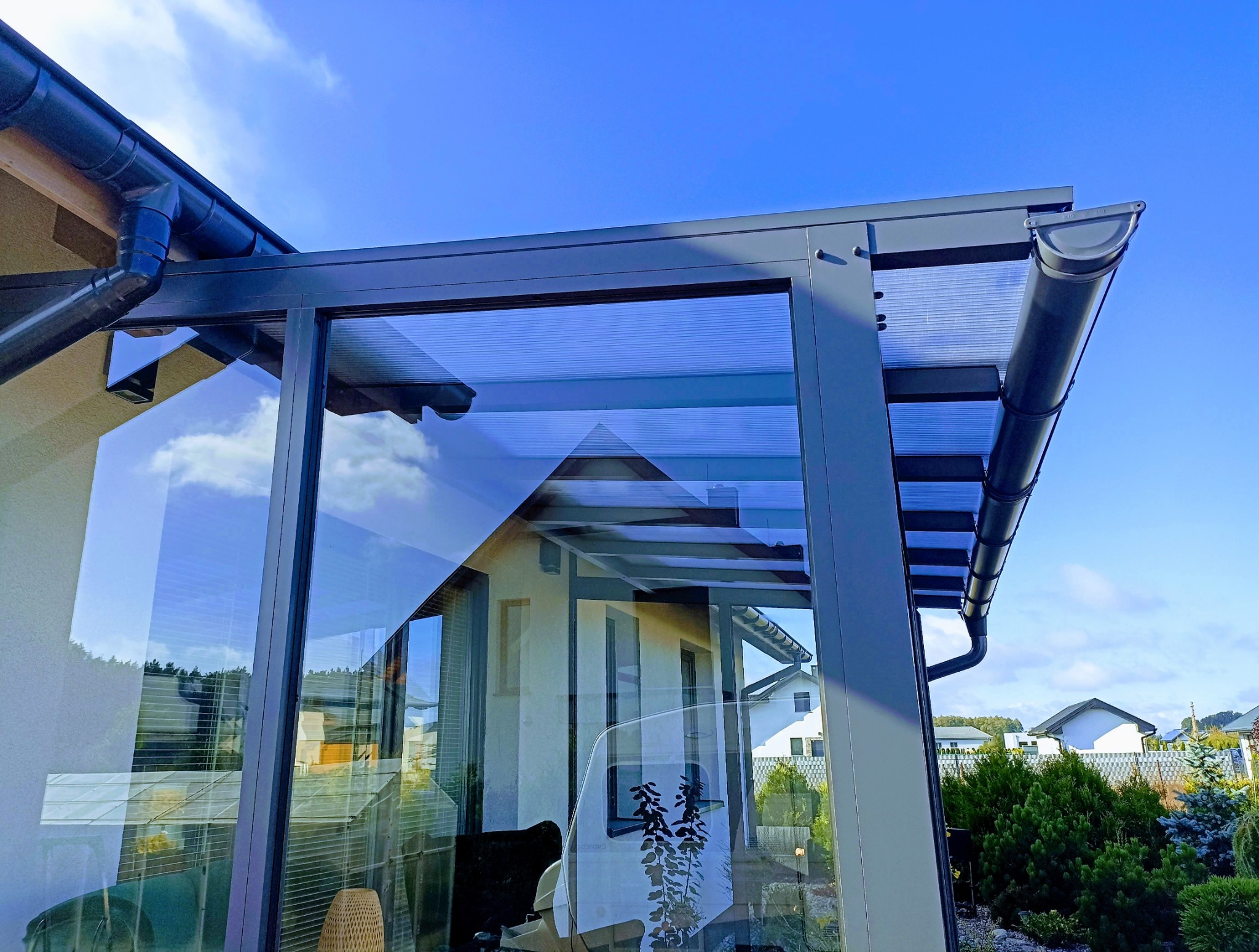 Szklana zabudowa balkonu z szarymi profilami i widocznymi rynnami, odbijająca otoczenie. Ujęcie z dołu, na tle błękitnego nieba.