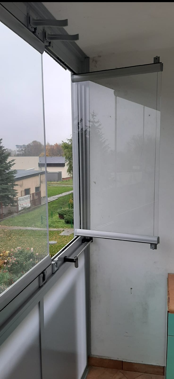 Otwarta, przesuwna zabudowa balkonu z profili aluminiowych i szkła. Widok na zewnątrz przez częściowo otwarte panele.