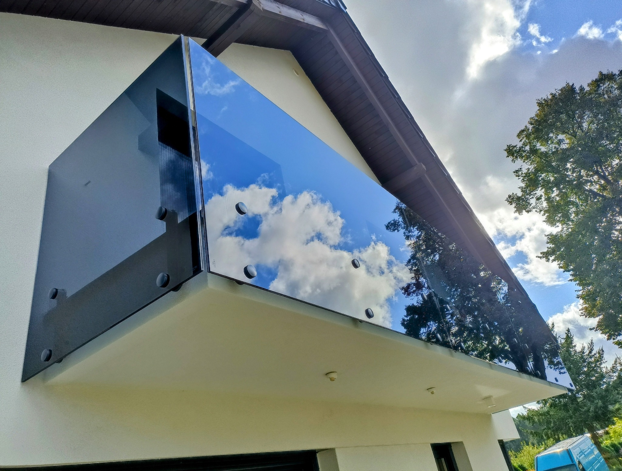 Szklana balustrada balkonowa z widocznym odbiciem chmur na szkle, zamocowana na jasnej elewacji budynku, z drzewem w tle.