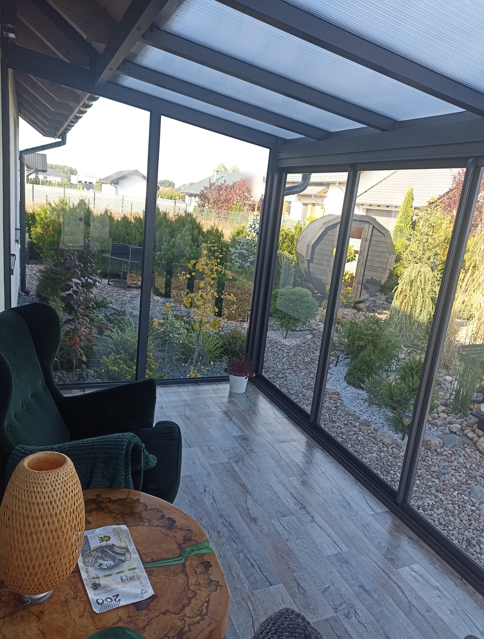 Nowoczesna zabudowa balkonu z czarnymi ramami, widok na ogród z drewnianą sauną. Wnętrze z fotelem i stolikiem z lampą.