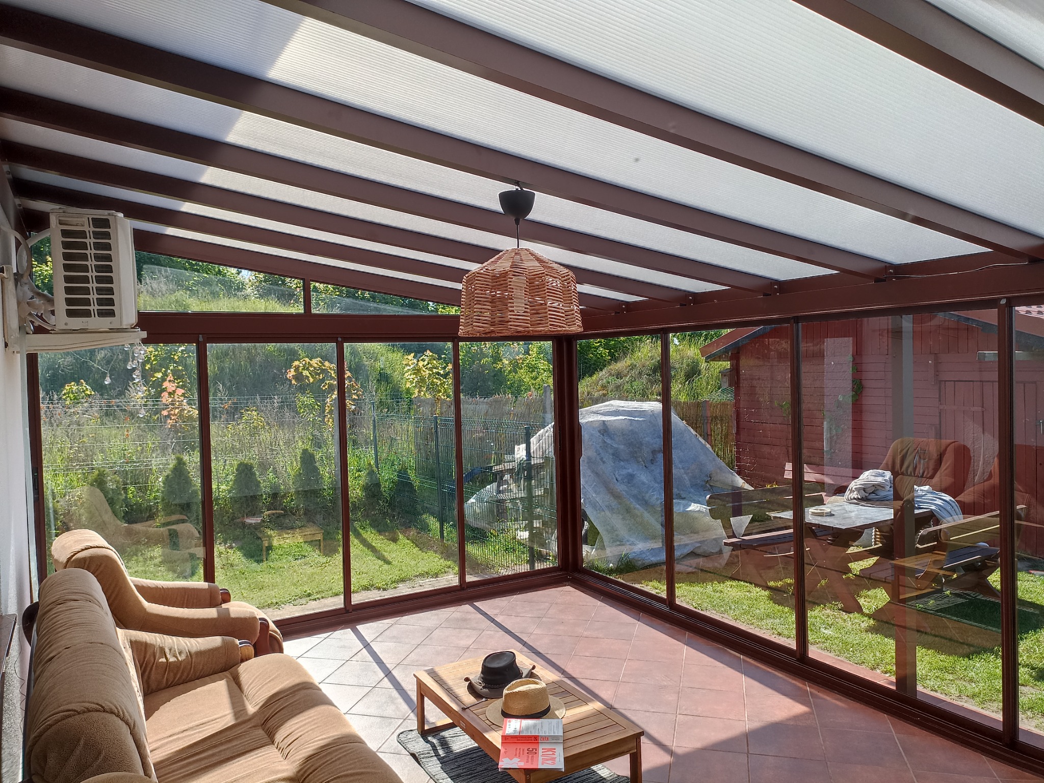 Zabudowany balkon z brązową ramą, widok na ogród. Wewnątrz sofa, stolik kawowy z kapeluszami i oświetlenie. Widoczny klimatyzator i przeszklona konstrukcja.