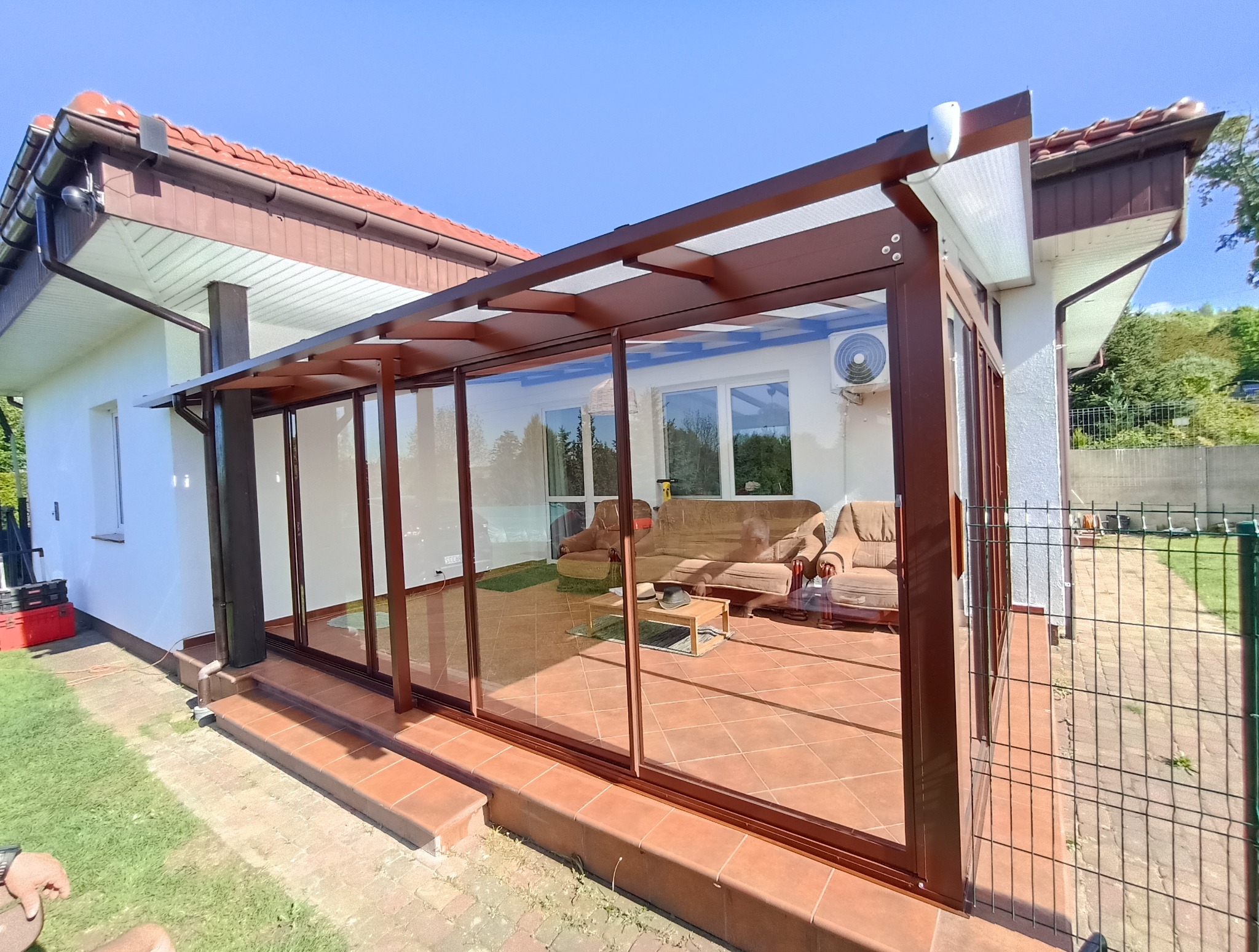 Zadaszony taras z brązową konstrukcją i szklanymi ścianami, widok na wnętrze z meblami wypoczynkowymi. Zewnętrzna część z trawnikiem i ogrodzeniem panelowym.