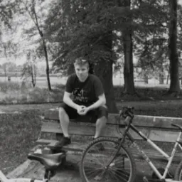 Czarno-białe zdjęcie mężczyzny siedzącego na ławce w parku, obok roweru górskiego. Twarz oświetlona, w tle drzewa i jezioro. Ujęcie z dołu.