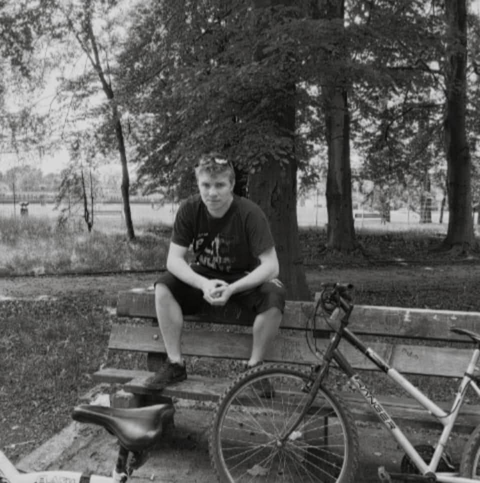 Czarno-białe zdjęcie mężczyzny siedzącego na ławce w parku, obok roweru górskiego. Twarz oświetlona, w tle drzewa i jezioro. Ujęcie z dołu.