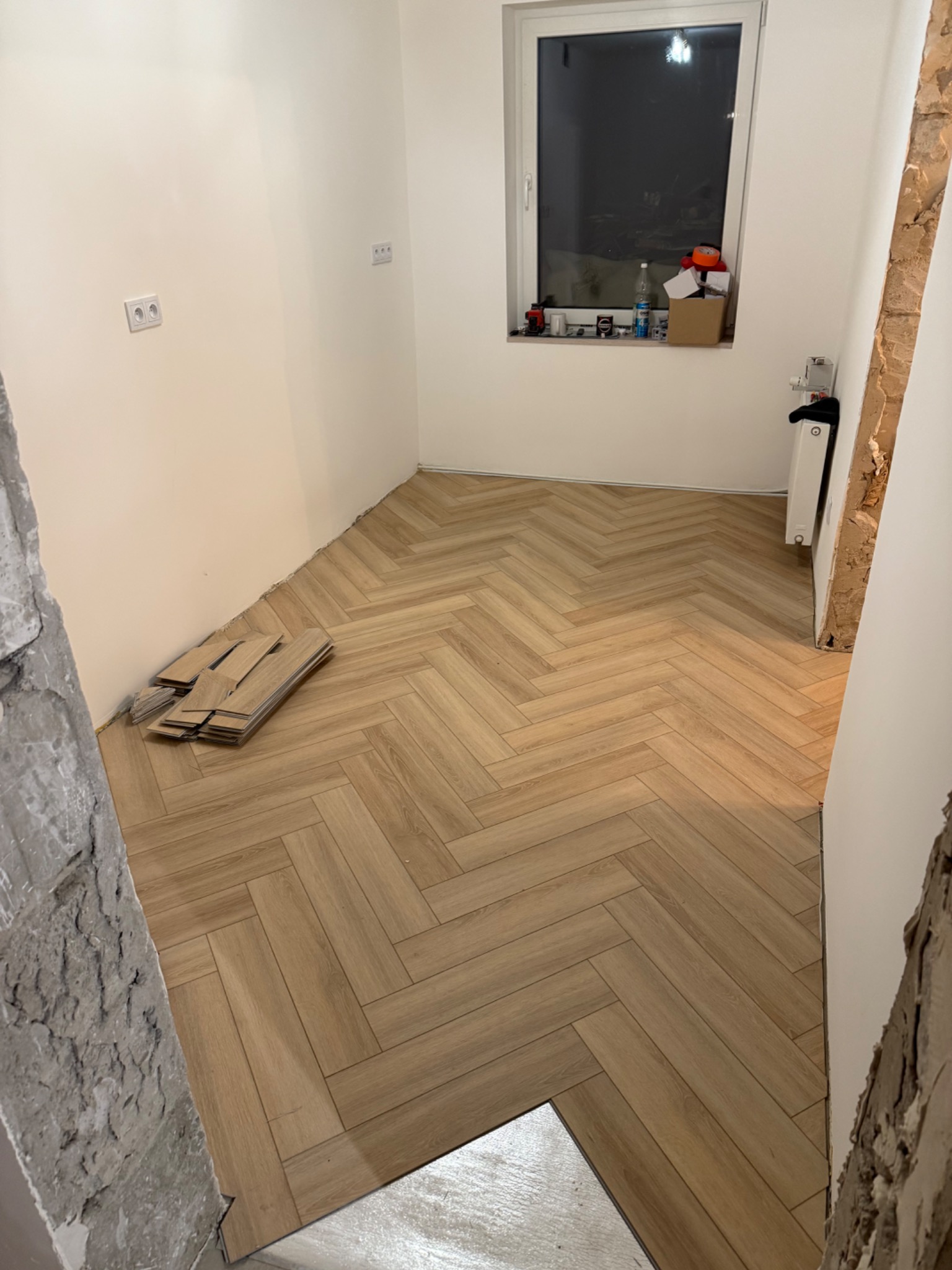 Ukończone układanie paneli podłogowych w jodełkę w jasnym pomieszczeniu. Na podłodze leżą jeszcze nieużyte panele. Fragment ściany w stanie surowym.