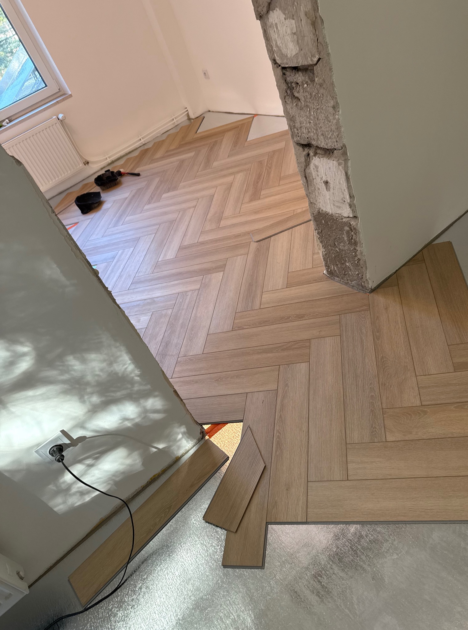 Układanie parkietu w jodełkę francuską, widoczny fragment ściany z gniazdkiem elektrycznym i narzędzia. Częściowo położona podłoga z panelami i fragment folii pod panele.