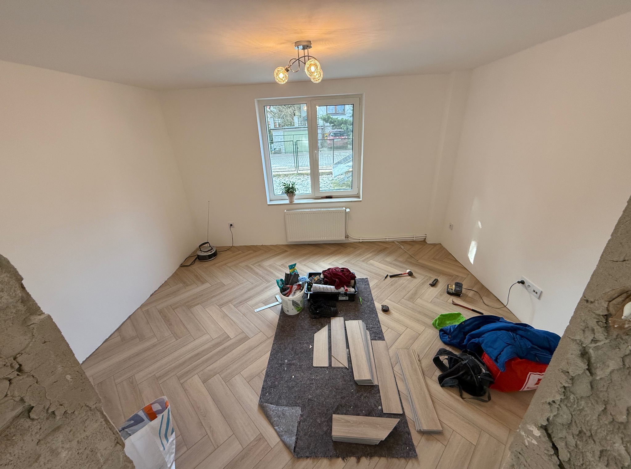 Układanie parkietu w jodełkę w jasnym pokoju z oknem. Widoczne narzędzia pracy i materiały. Częściowo ułożona podłoga, przygotowana do dalszych prac wykończeniowych.