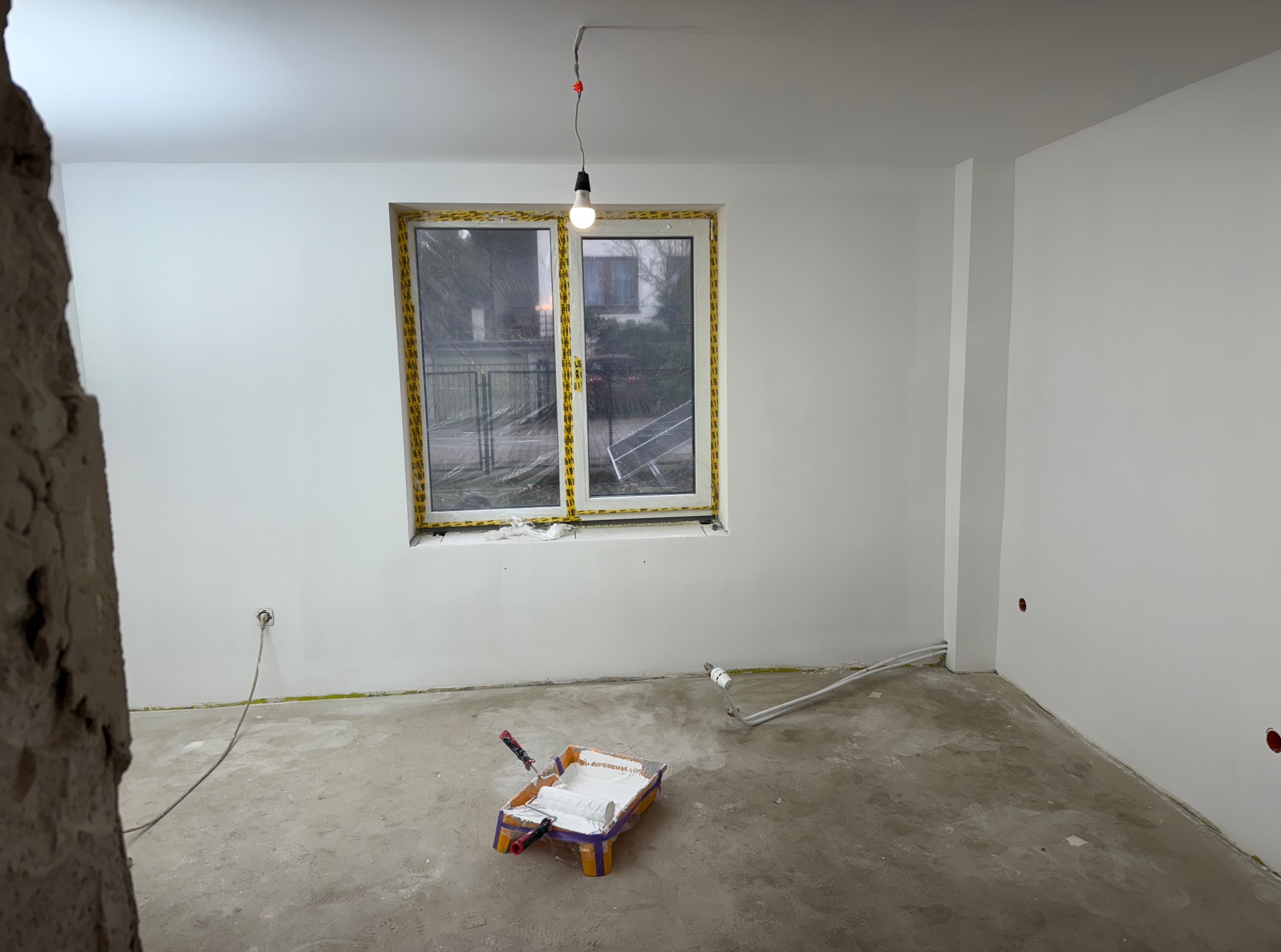 Białe, świeżo pomalowane ściany w pustym pokoju z oknem zabezpieczonym folią. Na podłodze kuweta z wałkiem malarskim. Widoczny fragment surowej ściany.
