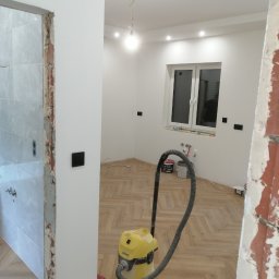 EBN-Bau - Remont wnętrza z parkietem ułożonym w jodełkę, widoczny odkurzacz Karcher, ściany po malowaniu, fragment łazienki w trakcie wykończenia i puszki z farbą.