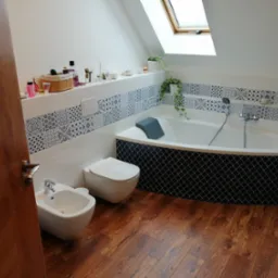 Jasna łazienka na poddaszu z drewnianą podłogą, białą armaturą i dekoracyjnymi płytkami. Uwagę przyciąga okno dachowe wpuszczające naturalne światło.