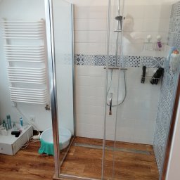 EBN-Bau - Wykończona łazienka z kabiną prysznicową typu walk-in, drewnopodobną podłogą i grzejnikiem drabinkowym. Widoczne białe płytki z dekoracyjnym pasem.