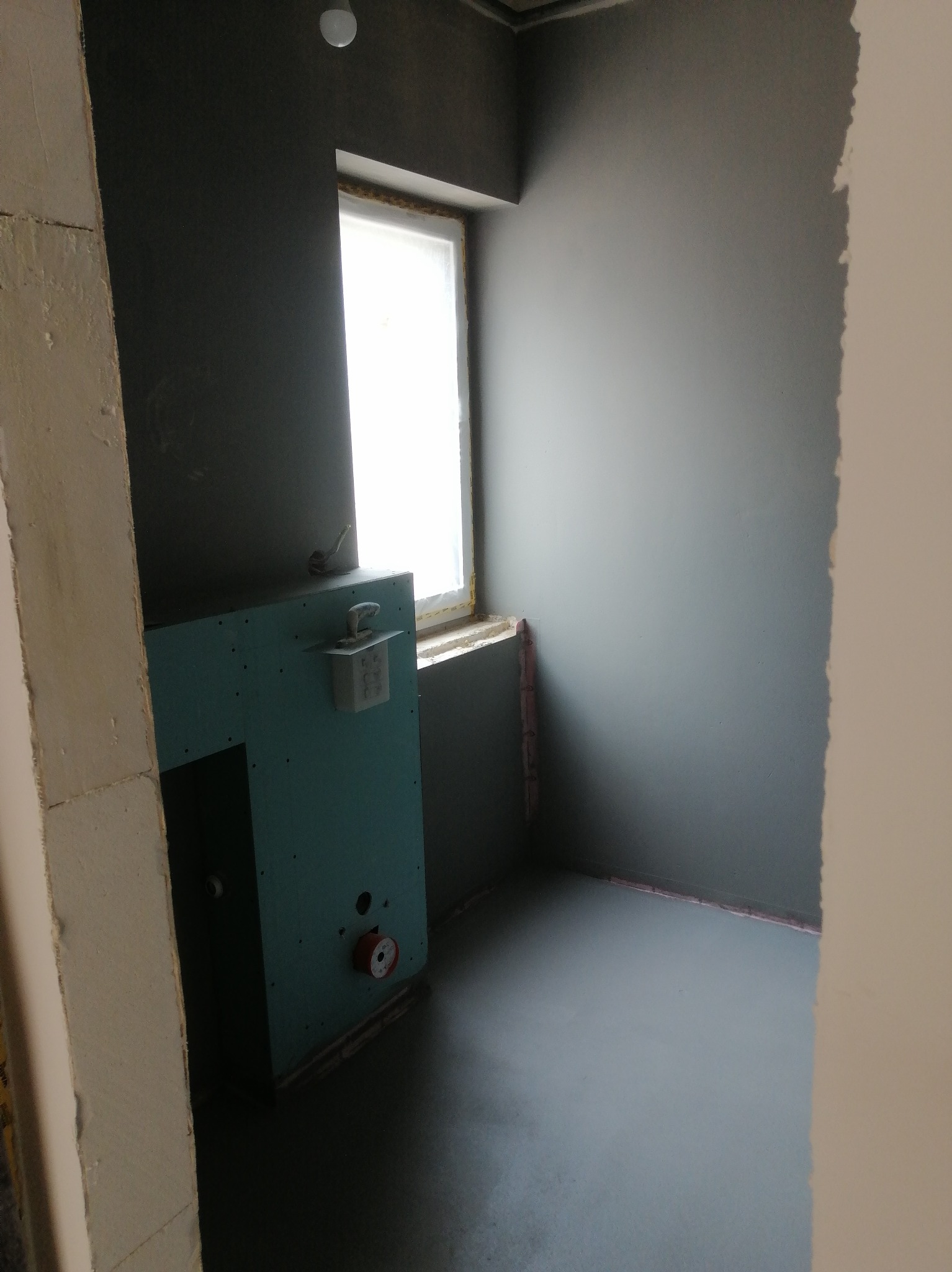 Łazienka w trakcie remontu: szare ściany, okno z uszczelnieniem, zabudowa z płyty GK z elementami instalacji wod-kan, wylewka podłogowa. Widok z perspektywy wejścia.