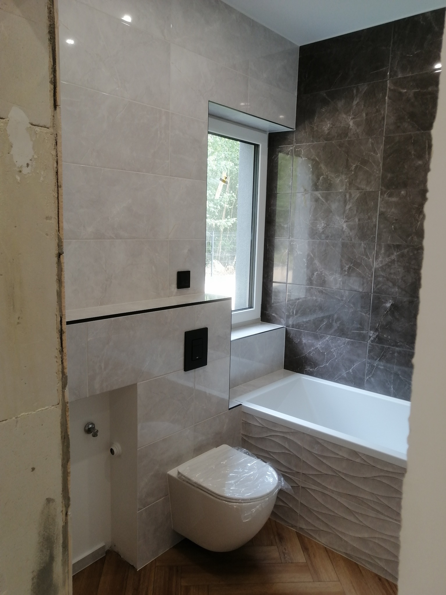 Wykończona łazienka z mozaiką, wiszącą toaletą i wanną z dekoracyjnym panelem. Widoczne płytki imitujące marmur i drewniana podłoga w jodełkę.