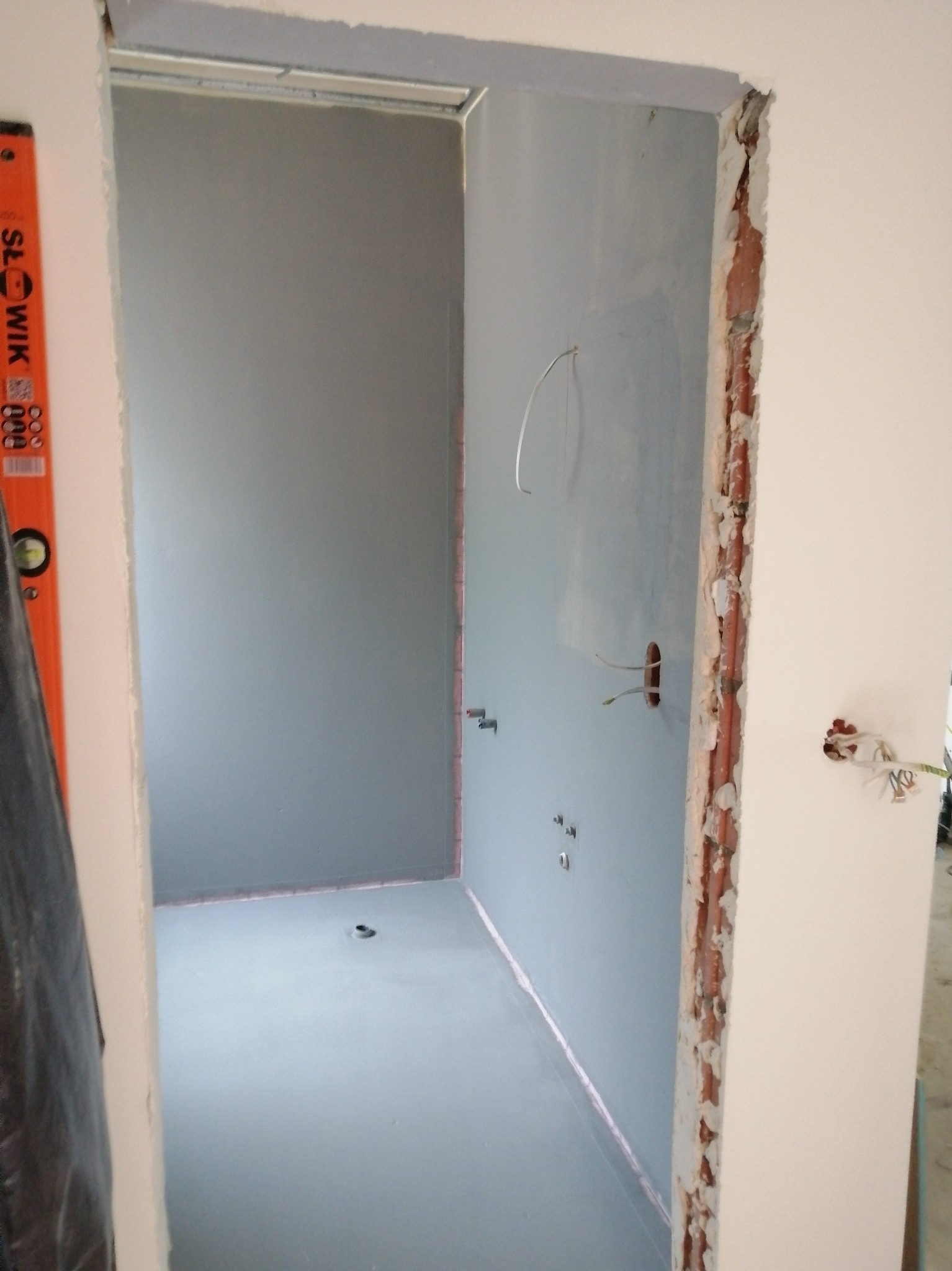 Wnętrze łazienki w trakcie remontu: ściany pokryte szarą farbą, widoczne instalacje, otwory na gniazdka i fragment poziomicy. Surowy stan wykończenia.