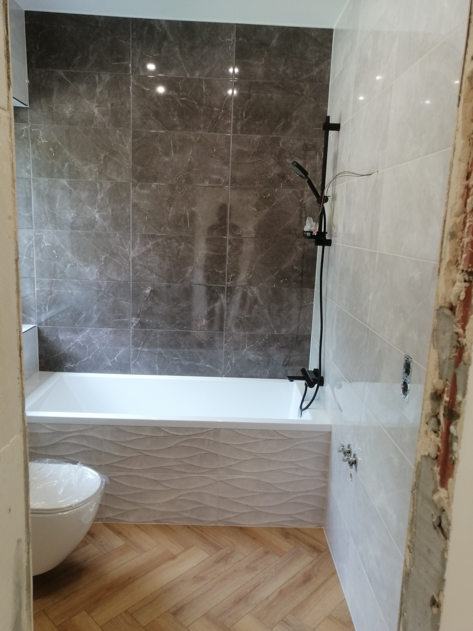Wykończona łazienka z wanną obudowaną płytkami o falistej strukturze, ciemnymi płytkami imitującymi marmur na ścianie i drewnopodobną podłogą. Widoczny czarny zestaw prysznicowy.
