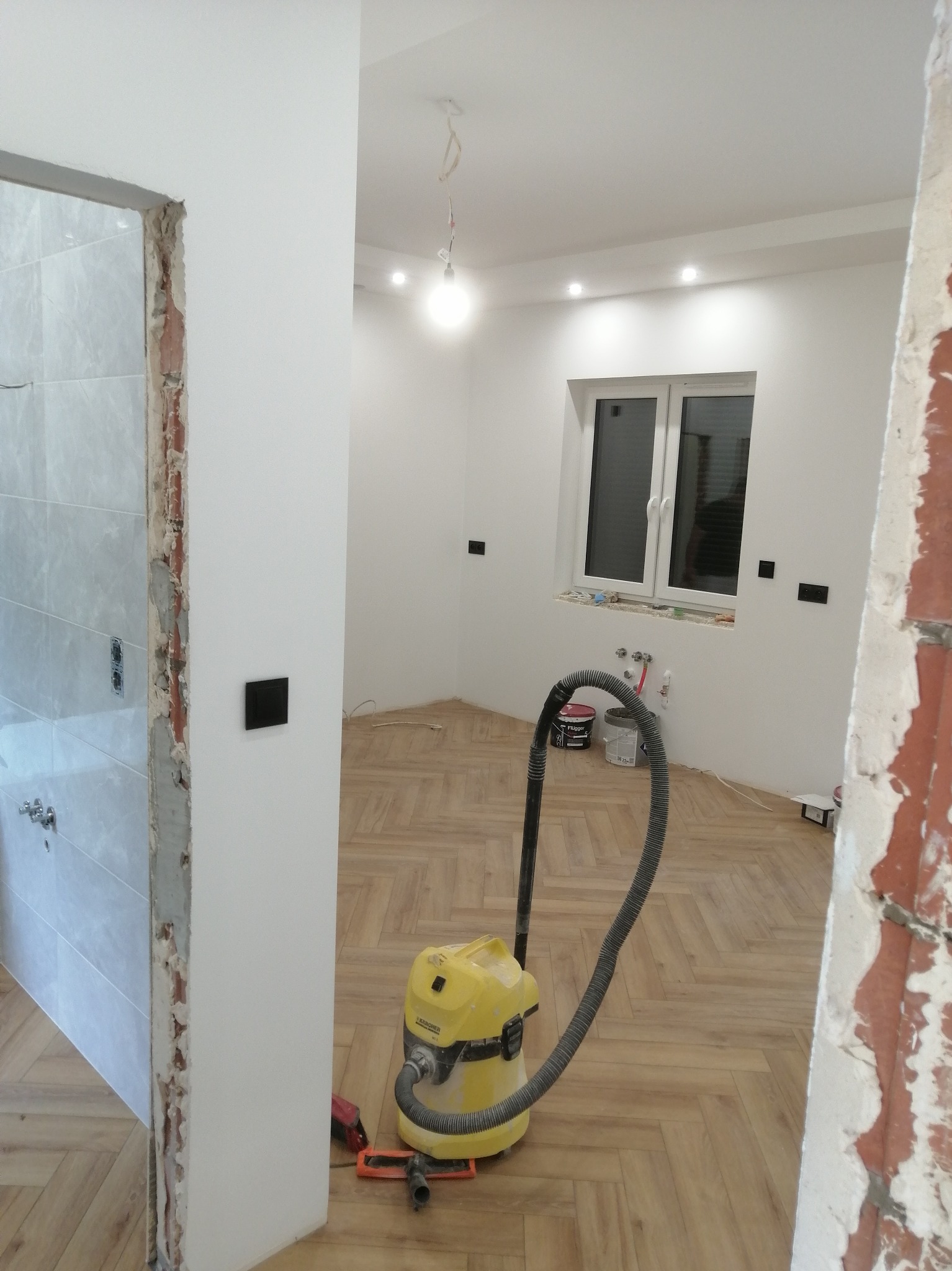 Remont wnętrza z parkietem ułożonym w jodełkę, widoczny odkurzacz Karcher, ściany po malowaniu, fragment łazienki w trakcie wykończenia i puszki z farbą.