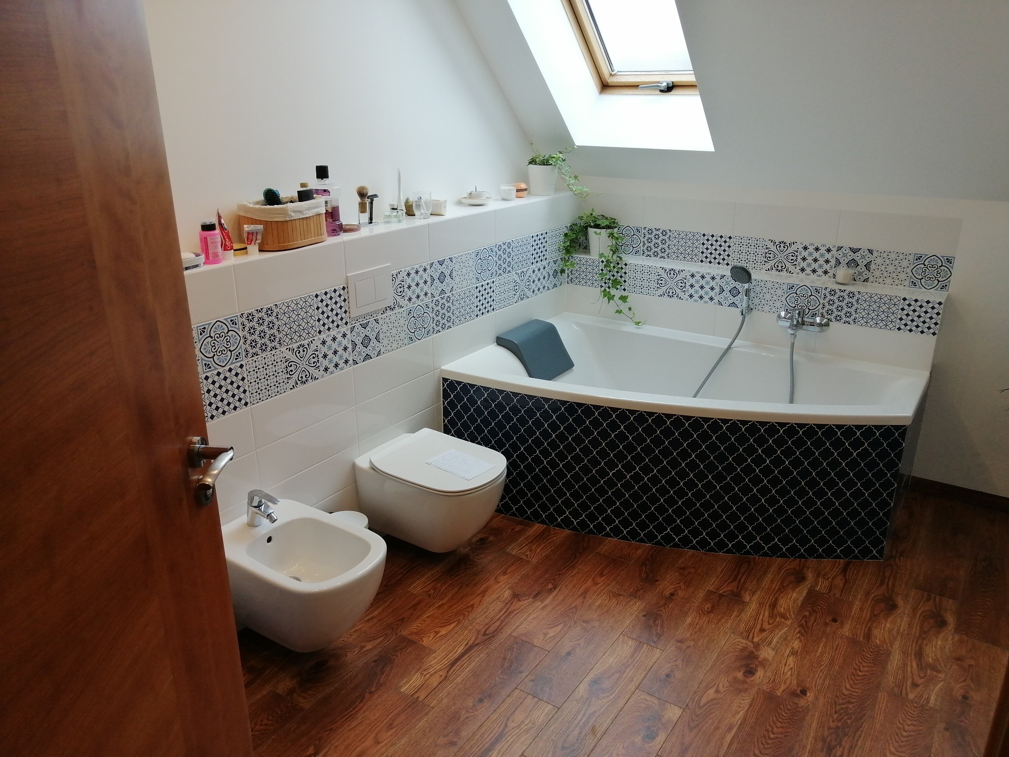 Jasna łazienka na poddaszu z drewnianą podłogą, białą armaturą i dekoracyjnymi płytkami. Uwagę przyciąga okno dachowe wpuszczające naturalne światło.