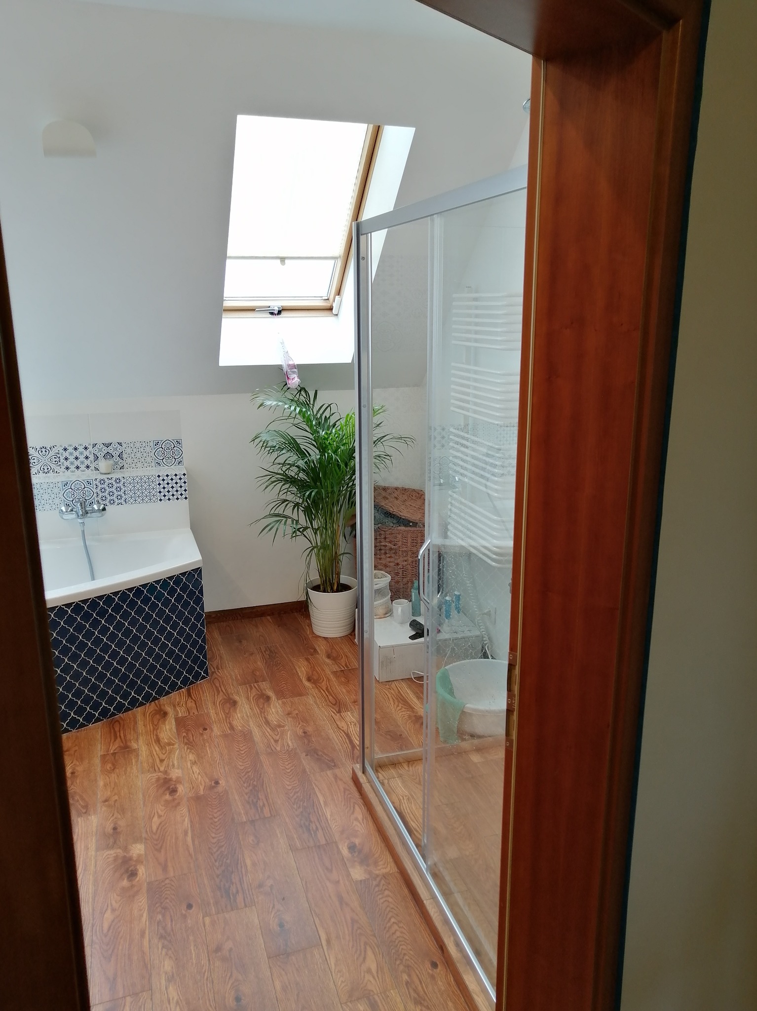 Jasna łazienka na poddaszu z drewnianą podłogą, kabiną prysznicową i oknem dachowym. Widok zza drewnianej framugi drzwi, z rośliną doniczkową w tle.