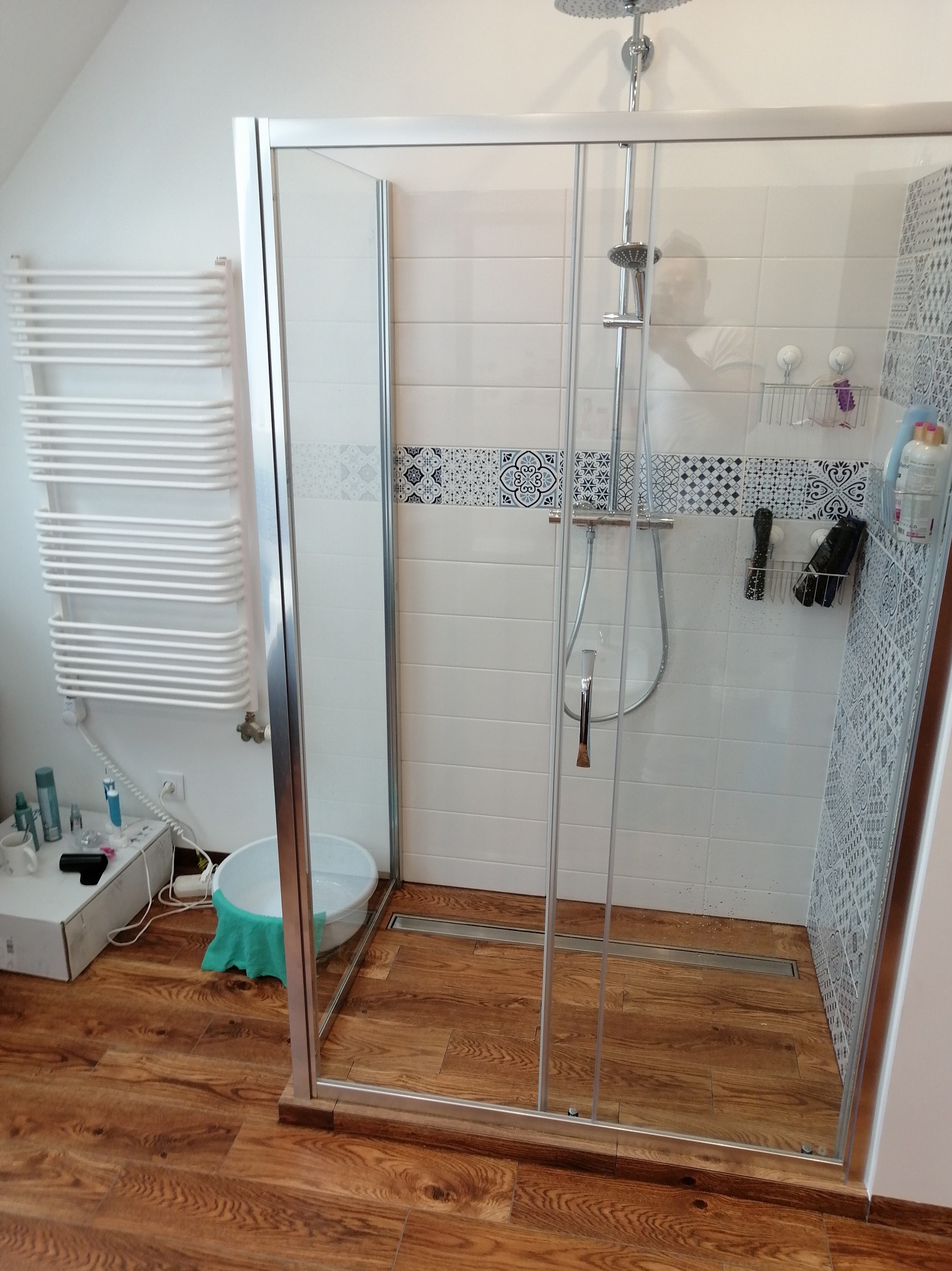 Wykończona łazienka z kabiną prysznicową typu walk-in, drewnopodobną podłogą i grzejnikiem drabinkowym. Widoczne białe płytki z dekoracyjnym pasem.