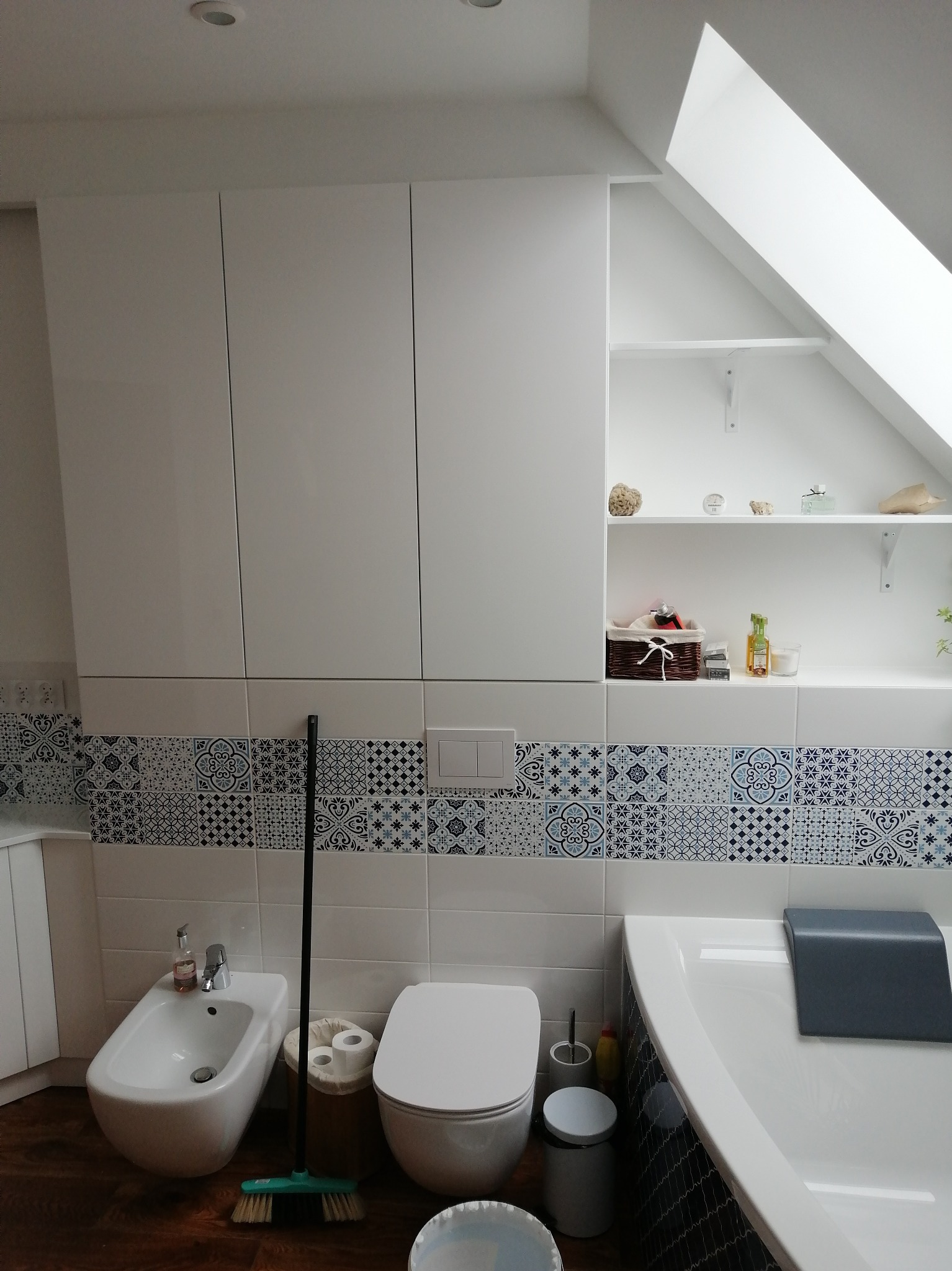 Biała łazienka na poddaszu z zabudową meblową, bidetem, toaletą i wanną. Płytki z motywem geometrycznym. Półki pod skosem z dekoracjami.