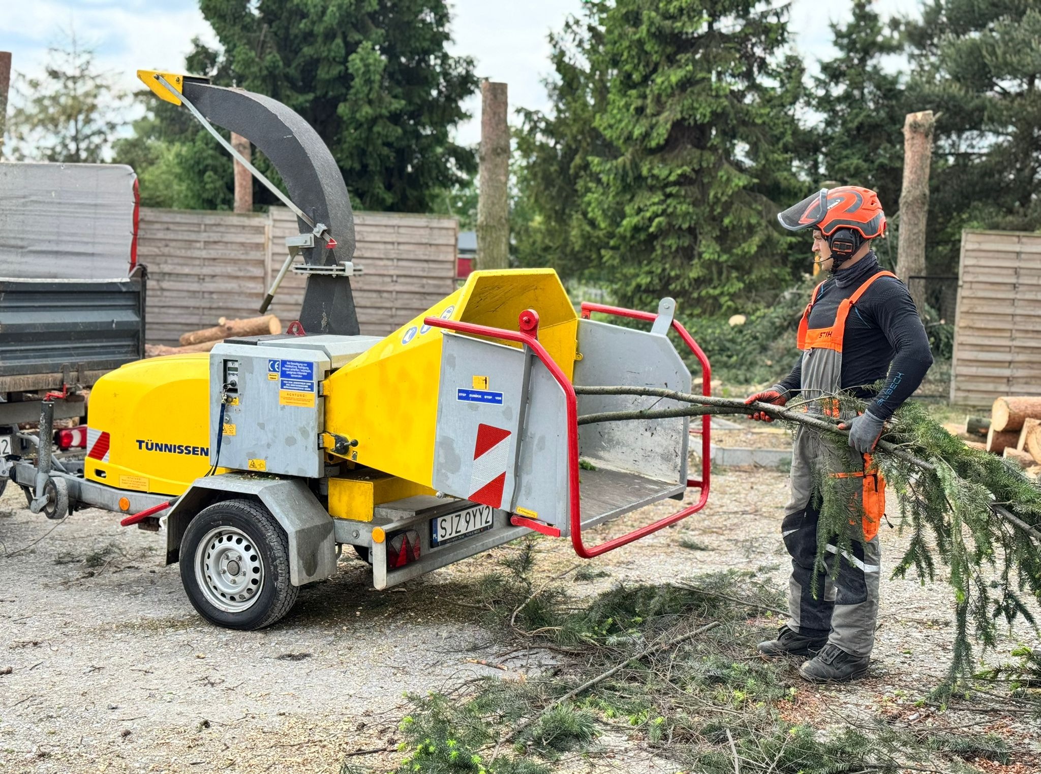 Pracownik w kasku i kombinezonie wrzuca gałęzie do rębaka Tünnissen. W tle ścięte drzewo i ogrodzenie.