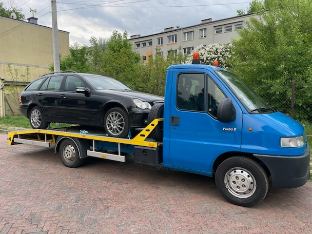 Niebieska laweta z czarnym kombi Mercedes na platformie, zaparkowana na brukowanej drodze, w tle budynek mieszkalny i zieleń.